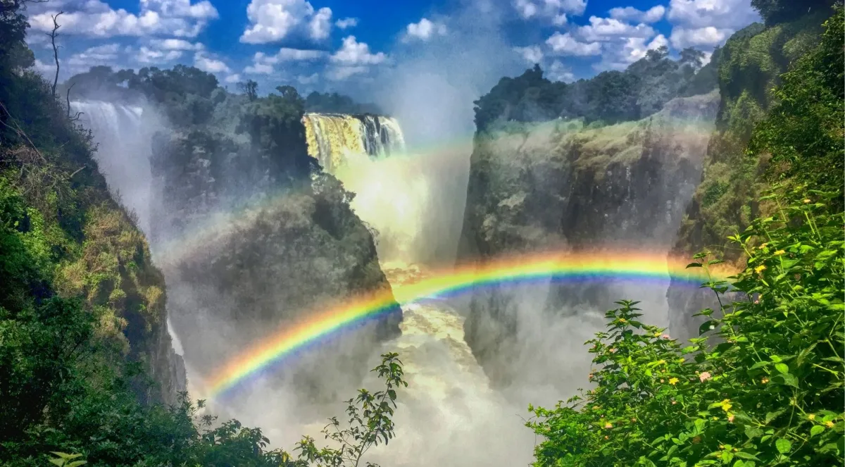A vivid double rainbow arcs over the spray of Victoria Falls, with lush tropical forest framing the cascade