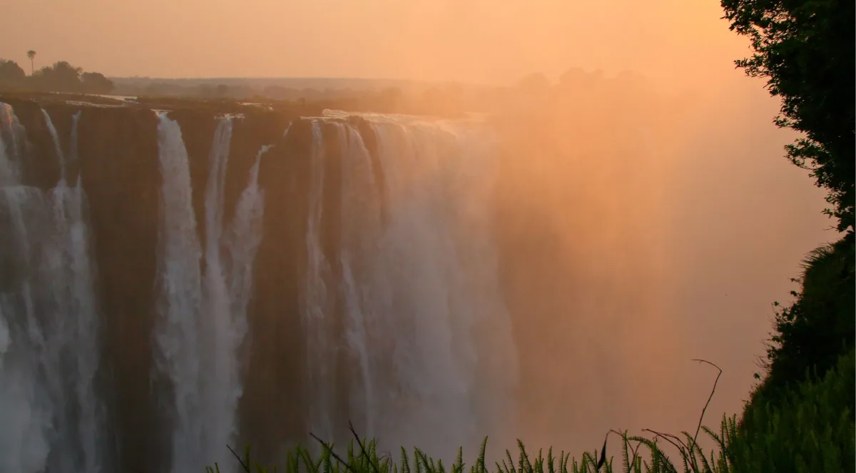 Victoria Falls glowing gold and amber at sunset, mist rising silently into a burnt-orange sky