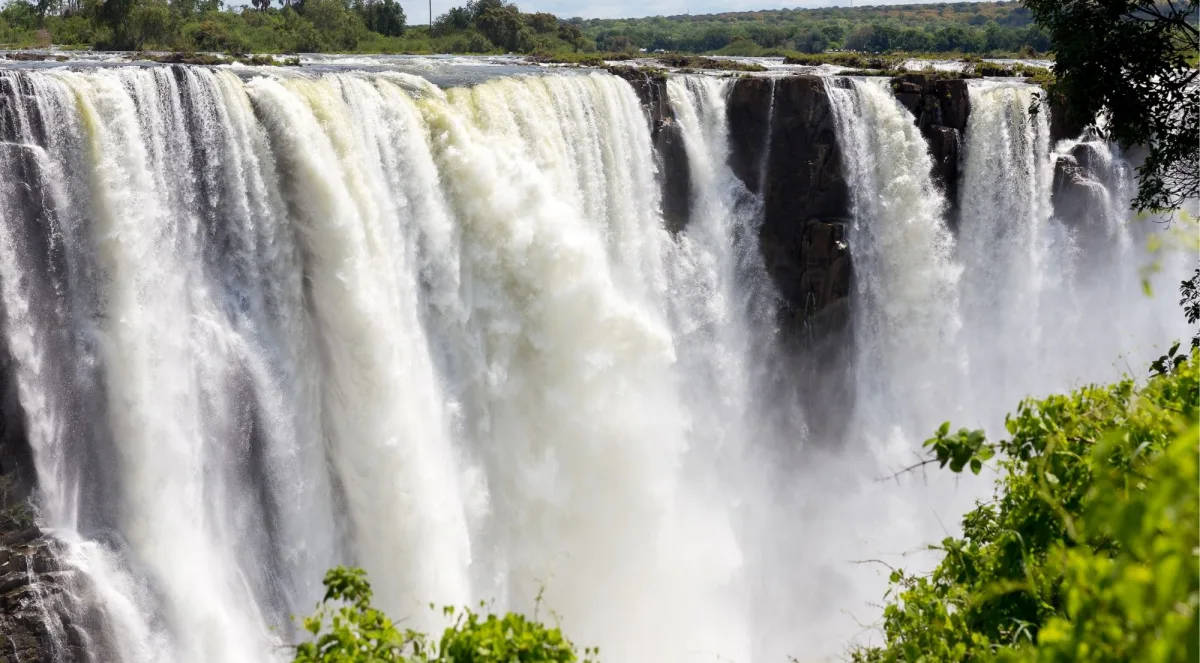 The full curtain of Victoria Falls thundering over its basalt lip, seen from across the gorge with green vegetation in the foreground