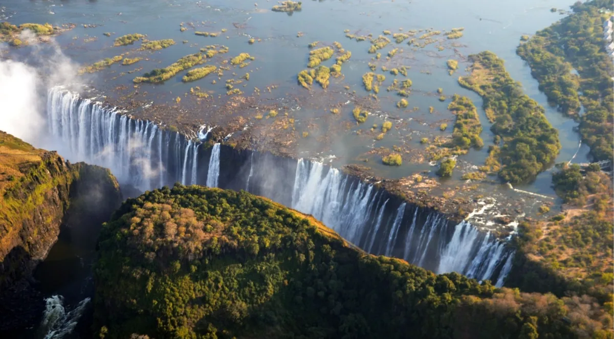 Aerial photograph of the upper Zambezi River spreading across forested islands just above Victoria Falls, the drop-off edge visible below